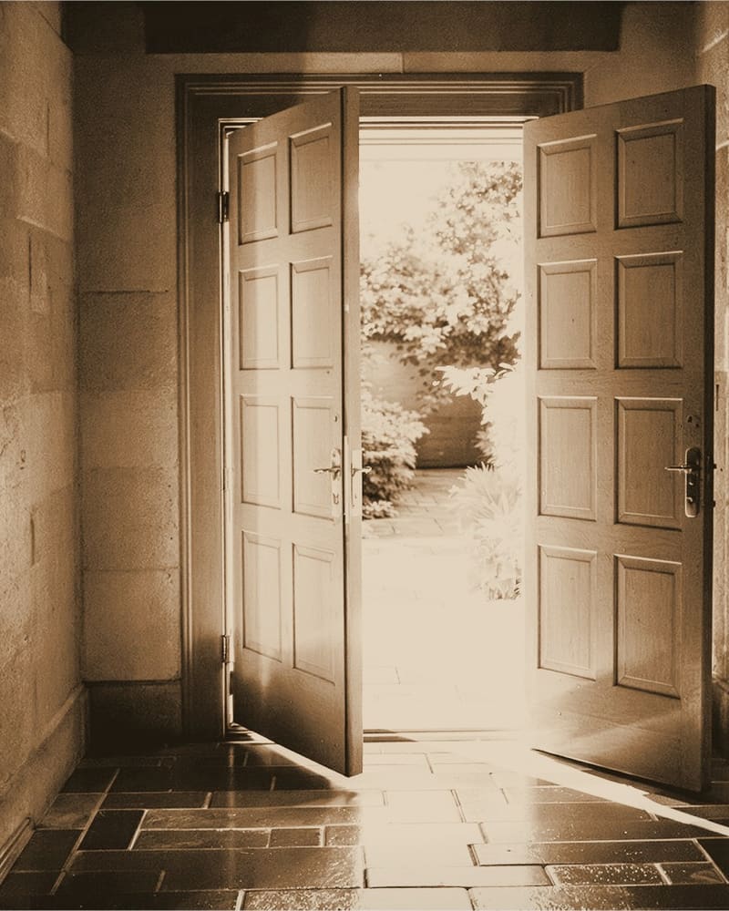 A pair of heavy panelled doors standing open onto a sunlit garden, warm light falling across stone flags.