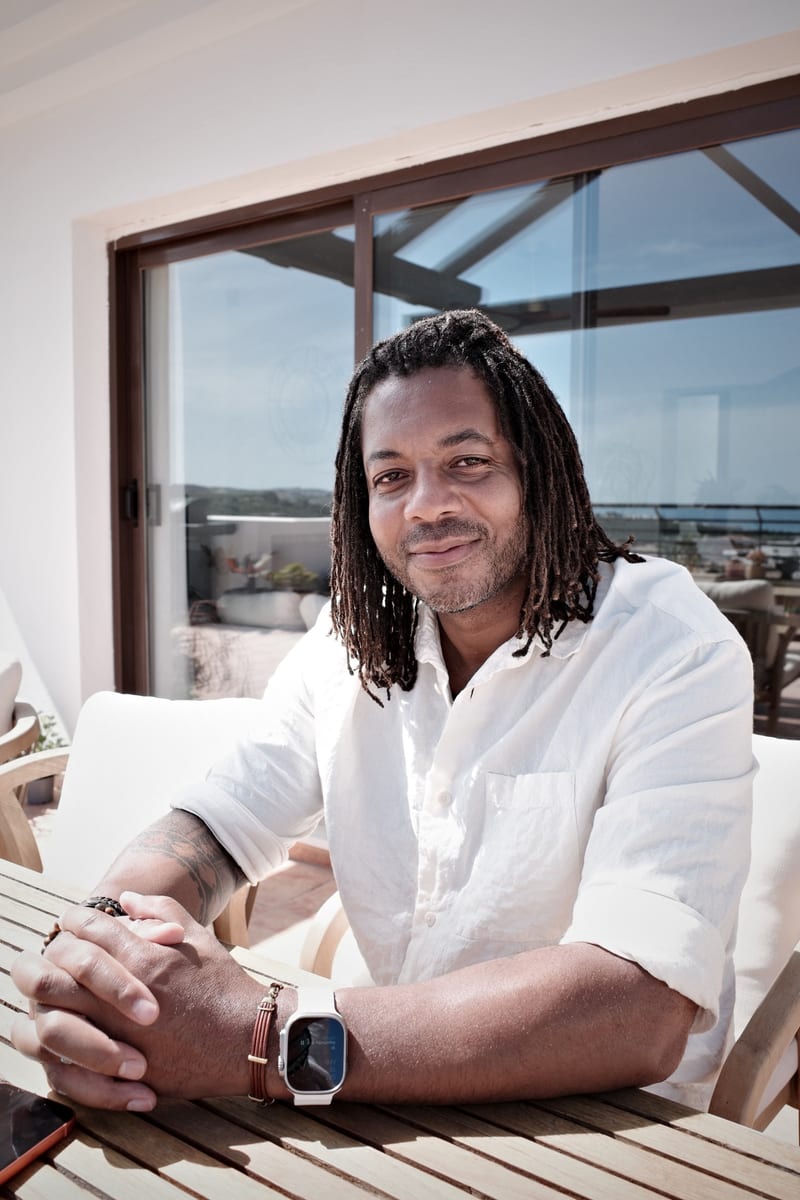 Nathan Lawes, smiling, in a white linen shirt at a table on a sunny terrace, with the hills and sea reflected in the glass door beside him